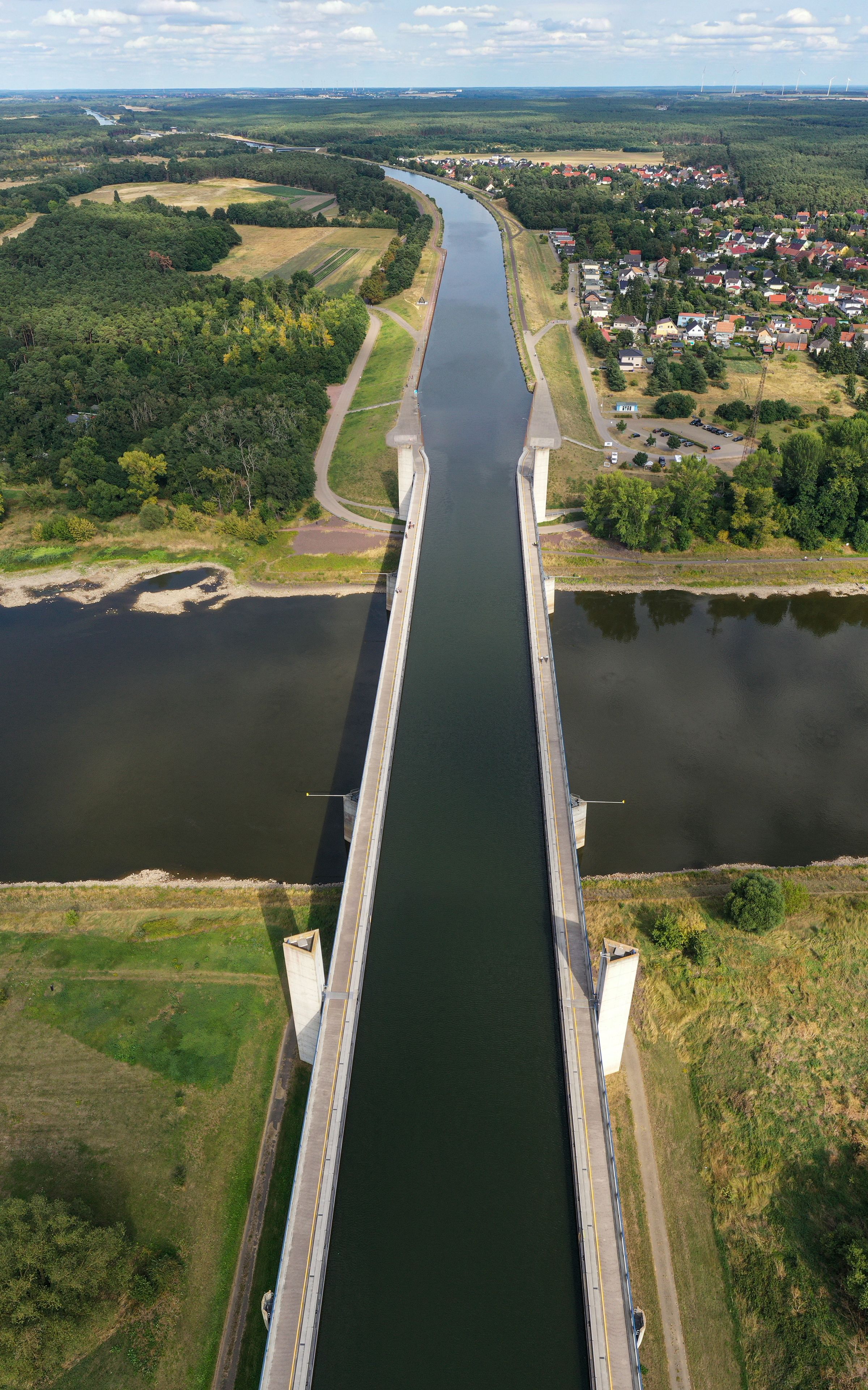 Luchtbeeld van een lange brug over een rivier, omringd door groene velden en een klein stadje met rode daken.