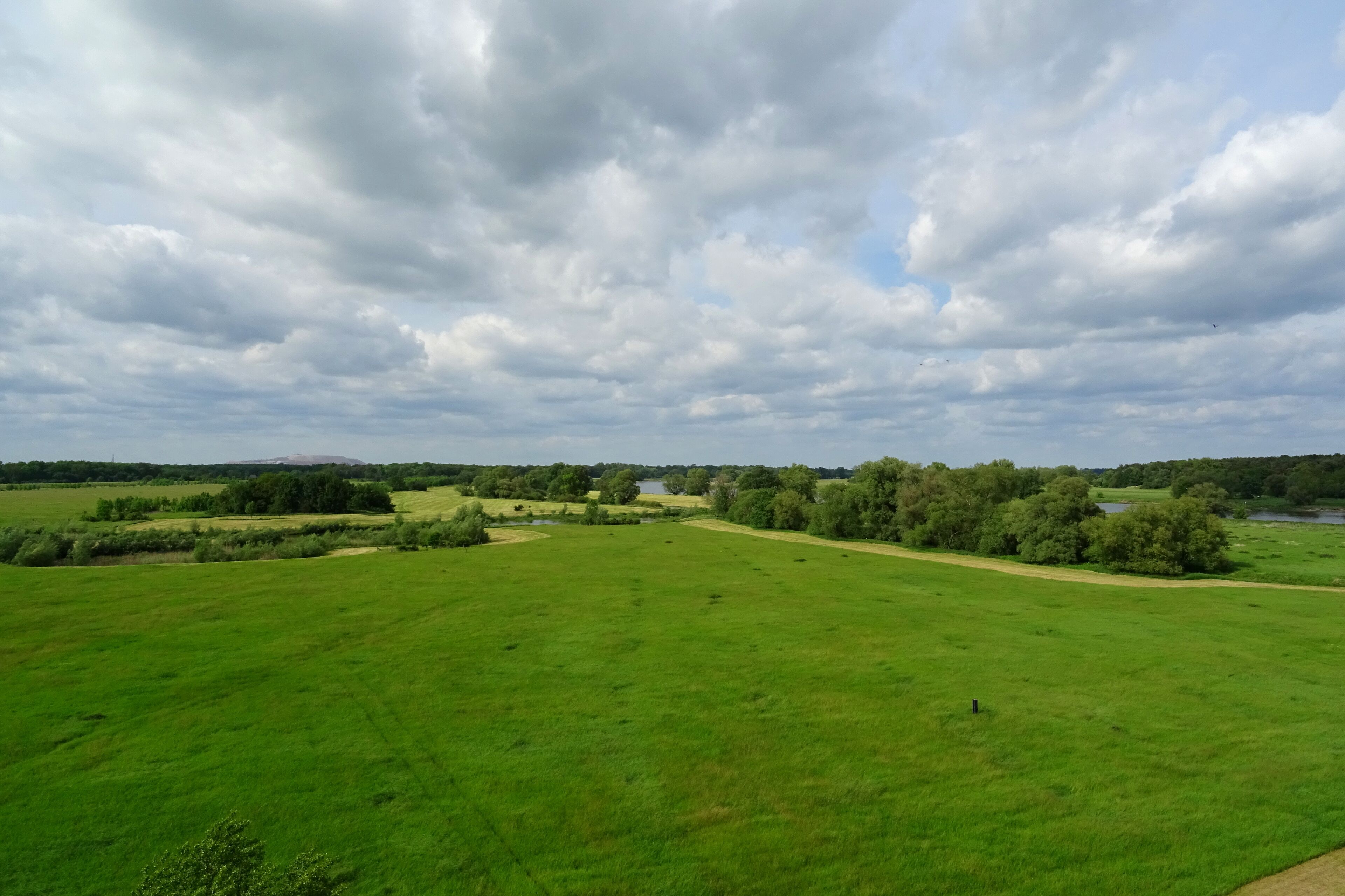 A vast green field with a river and trees in the distance under a cloudy sky.