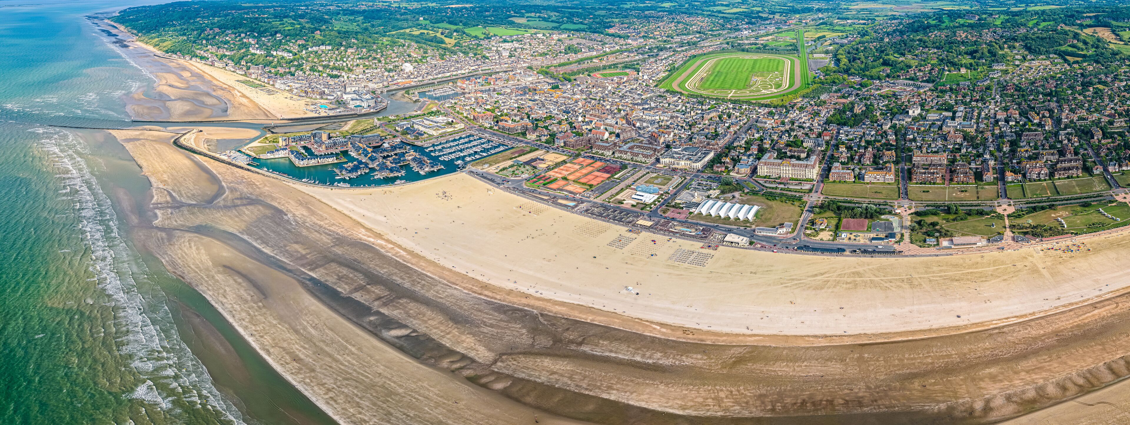 An aerial view of a coastal city with a marina, sandy beaches, and a large green sports field.