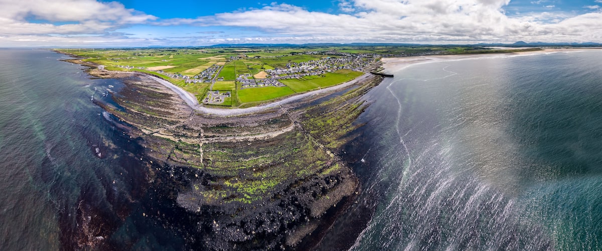 An aerial view of a coastal town with a harbor, green fields, and a clear sky.