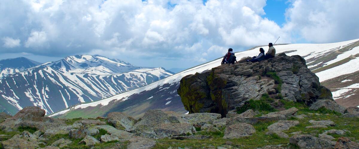 A rocky outcrop with two hikers, snow-capped mountains, and a clear sky.