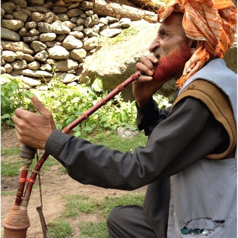 Una persona tocando una flauta tradicional al aire libre, con un muro de piedra y vegetación de fondo.