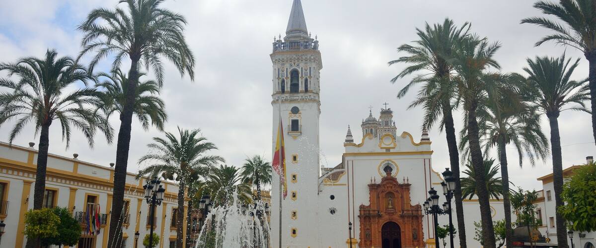 A historic plaza with a central fountain, palm trees, and a prominent church with a bell tower.
