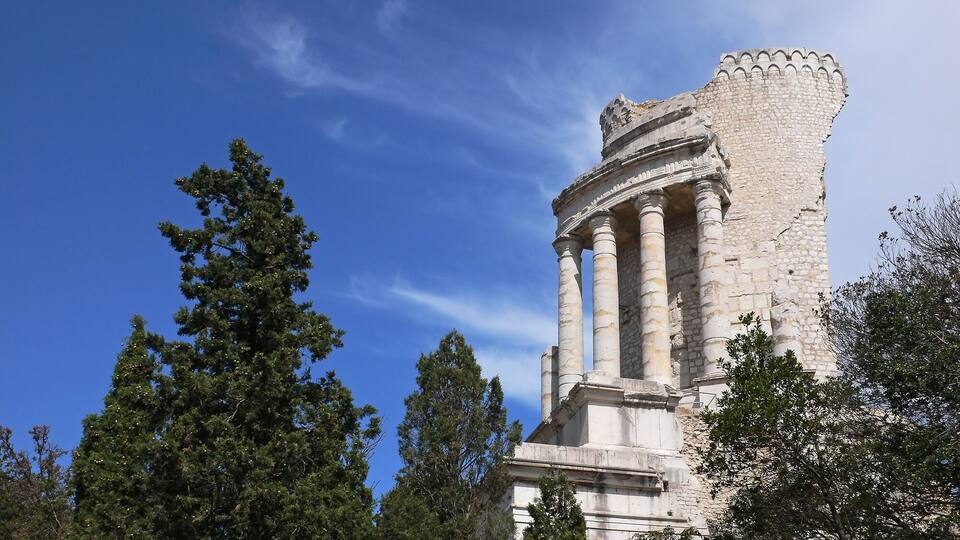 A historical stone structure with columns and a crenellated top, surrounded by trees.