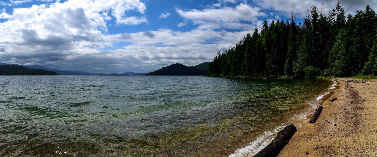 A beach with clear water, surrounded by dense forests and mountains in the background.