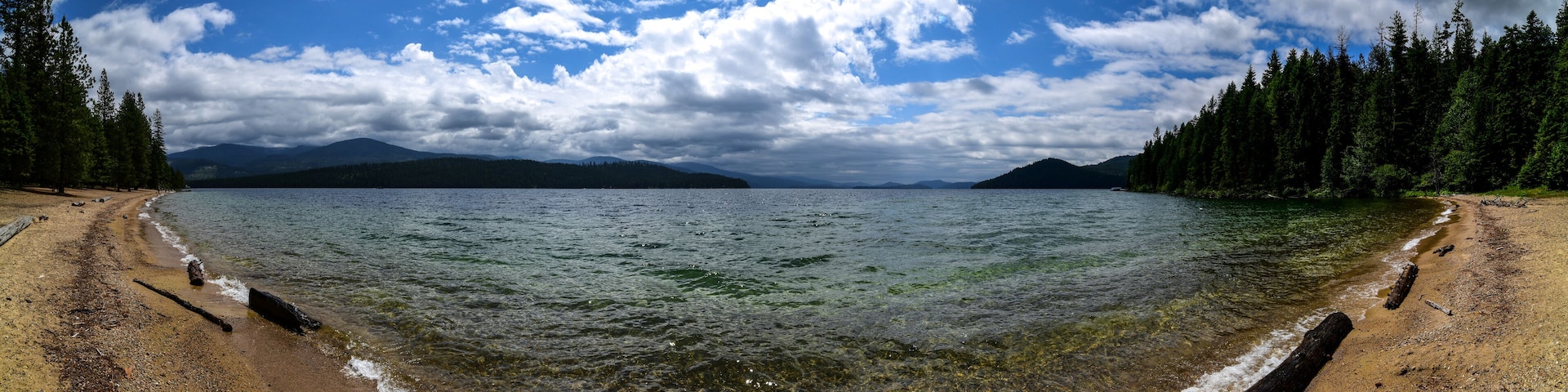 A beach with clear water, surrounded by dense forests and mountains in the background.