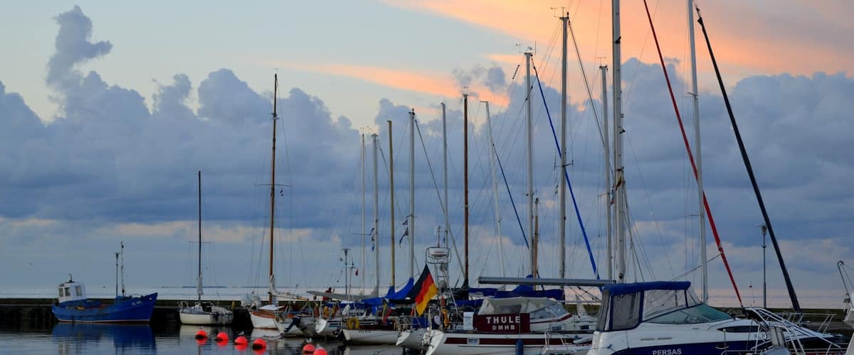 A marina with multiple sailboats docked, a German flag, and a sunset sky.