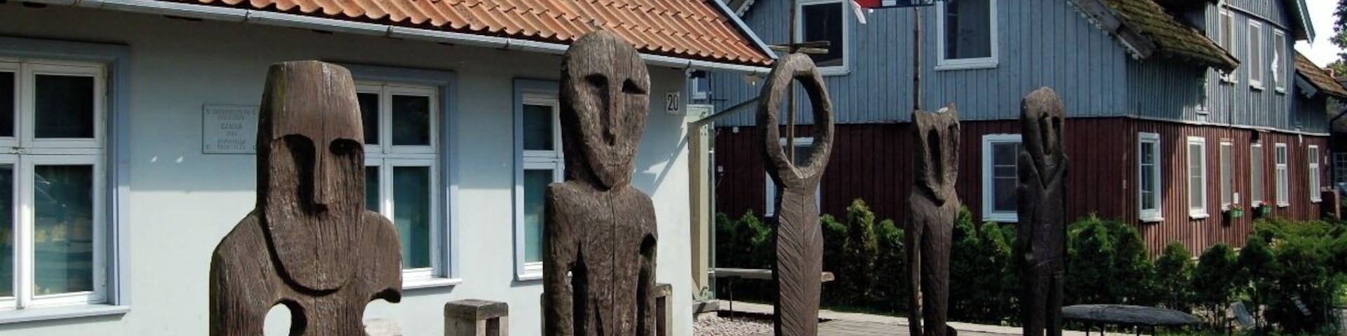 Esculturas de madeira em formato de figuras humanas em um jardim, com casas ao fundo.