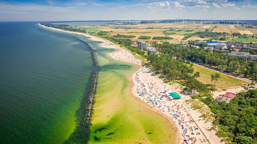 Une plage côtière aux eaux limpides, avec un rivage sablonneux et des éoliennes visibles au loin.
