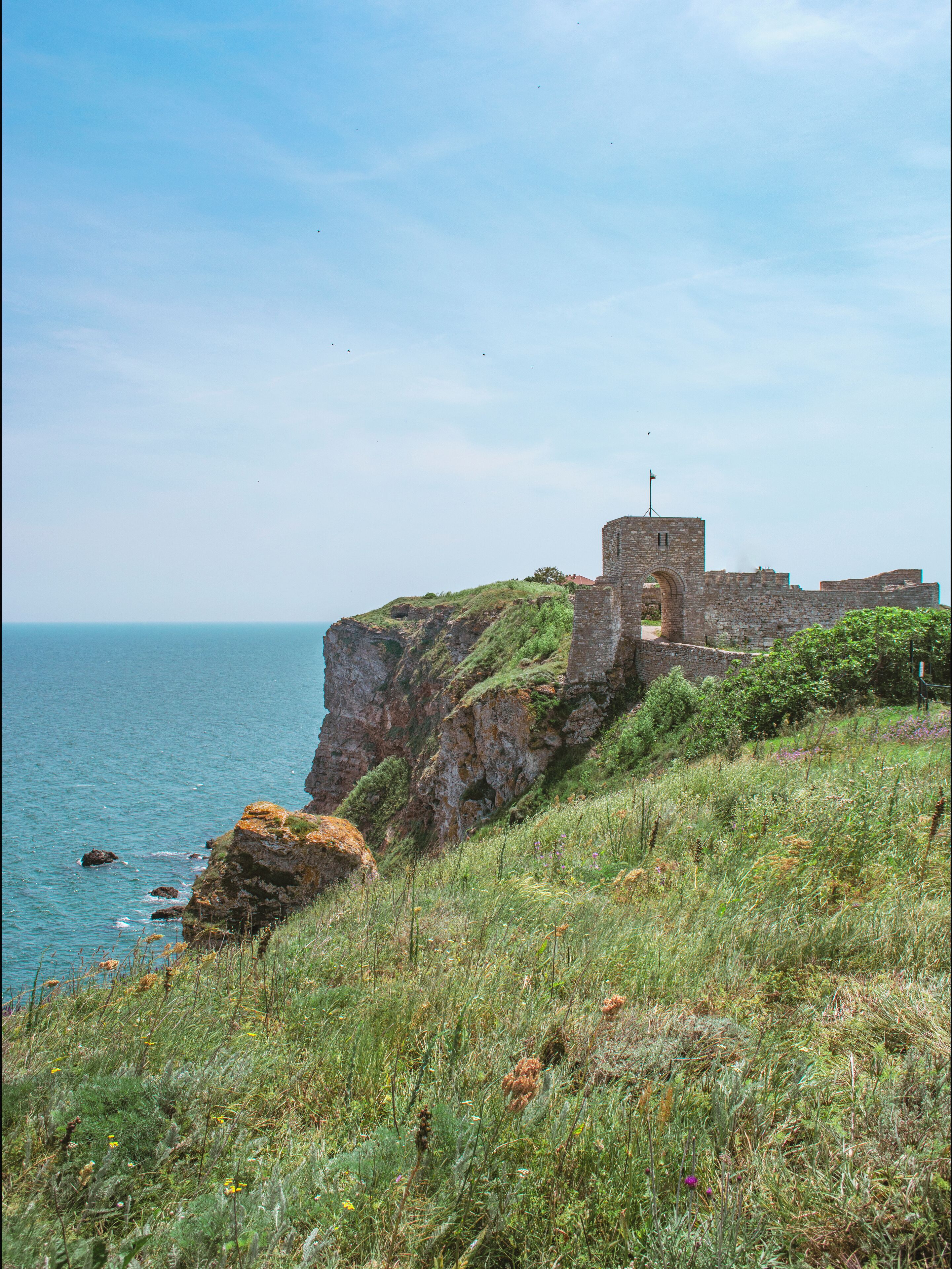 A coastal stone fortification with a tower, surrounded by grassy terrain and overlooking the sea.