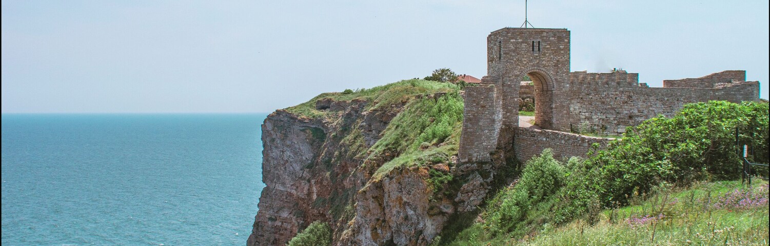 Uma fortificação costeira de pedra com uma torre, rodeada por terreno gramado e com vista para o mar.