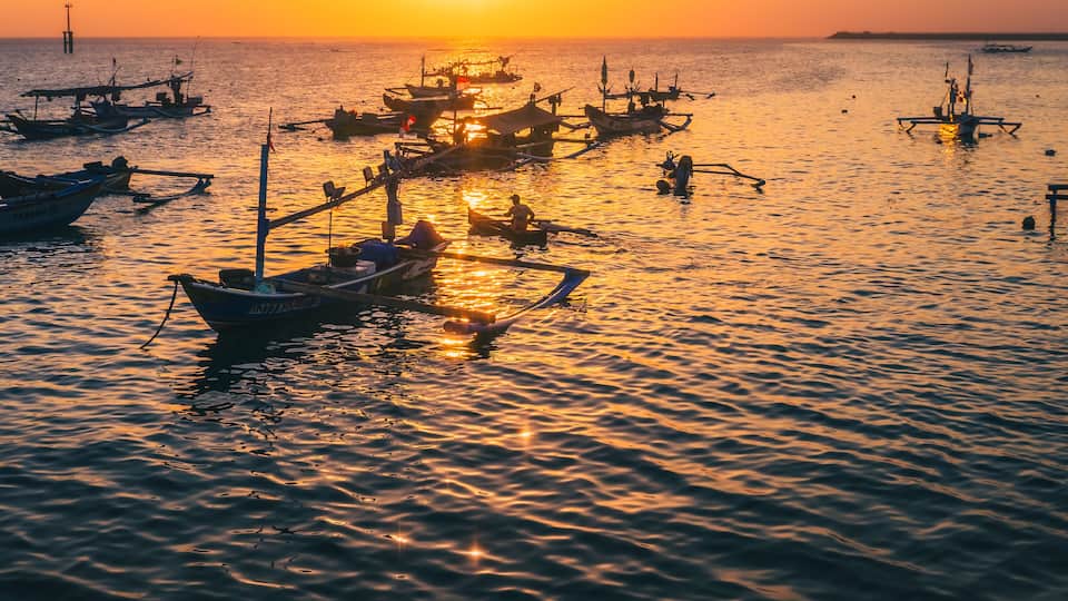 A sunset over a calm sea with several boats anchored, reflecting the warm colors of the sky.