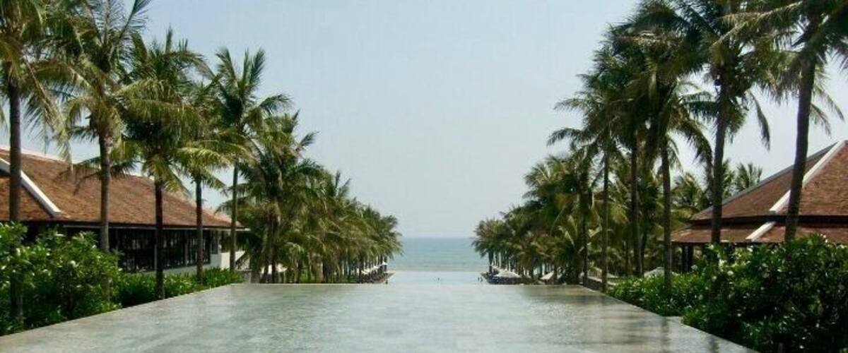 A long, empty pool with palm trees and overwater bungalows.