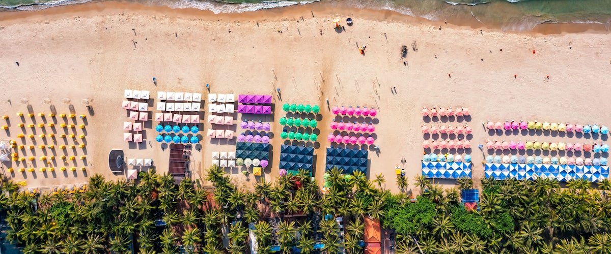 A beach with rows of colorful umbrellas and chairs, palm trees, and waves.