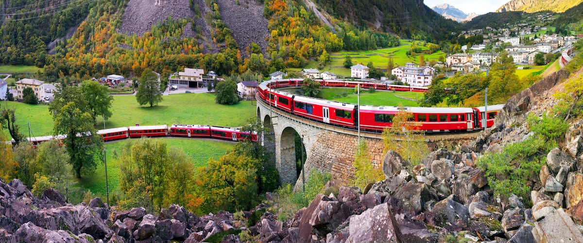 A red train traveling over a stone bridge in a mountainous area with houses and autumn foliage.