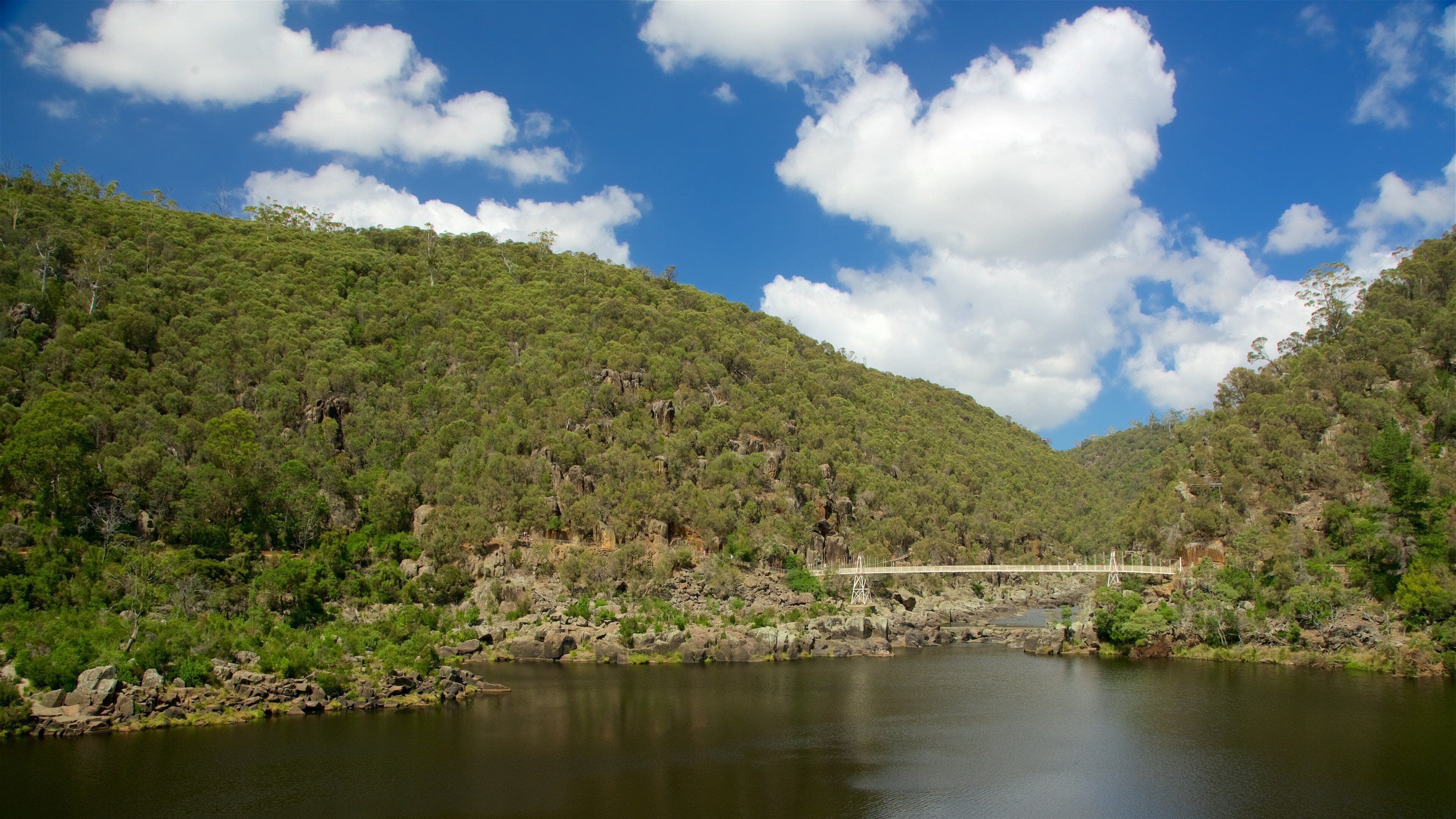 A river with a bridge, surrounded by green hills and a clear blue sky.