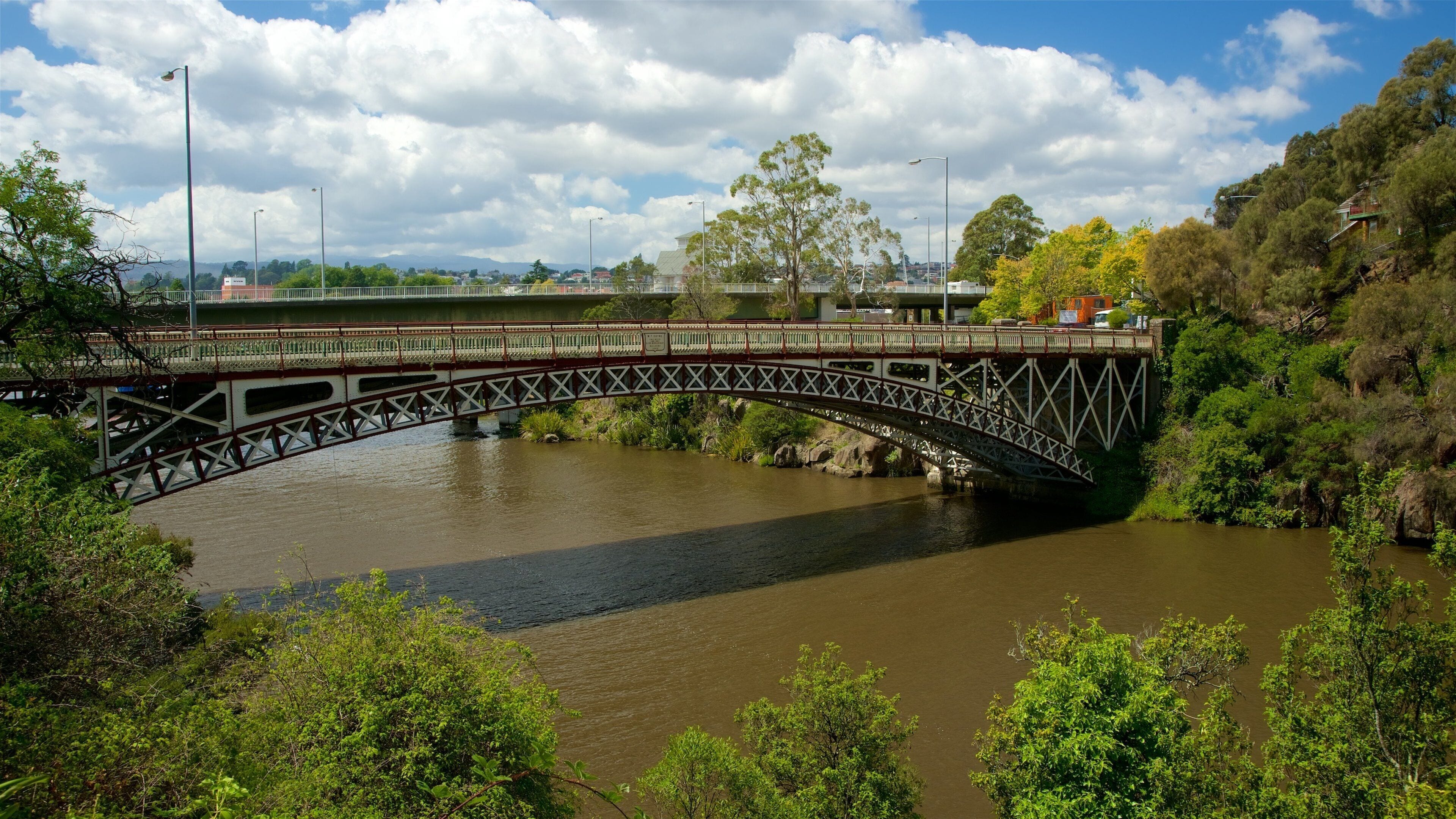 Un puente metálico sobre un río, con árboles y edificios al fondo.