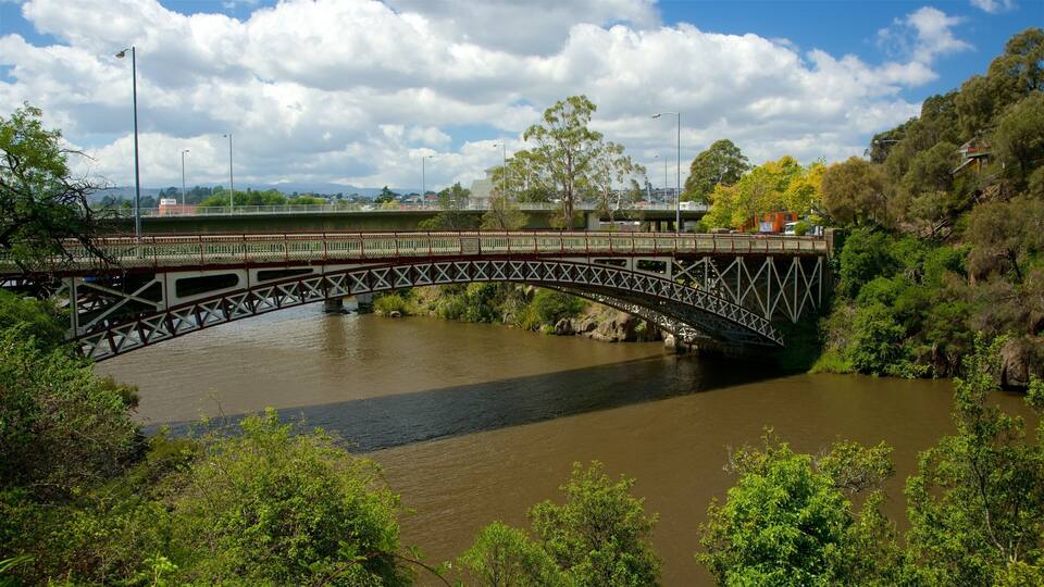Un puente metálico sobre un río, con árboles y edificios al fondo.