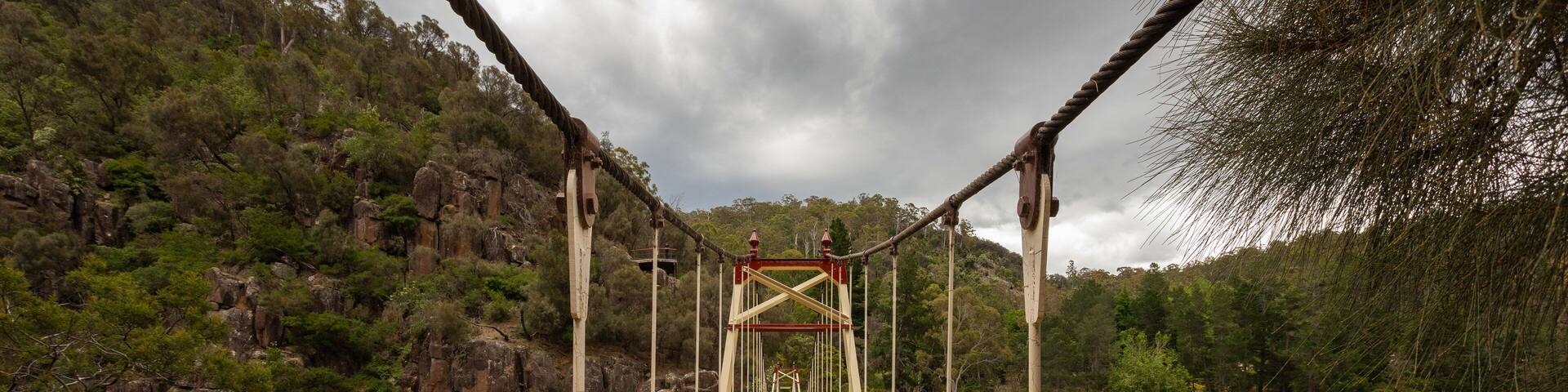 Un puente colgante sobre un río, con una ladera boscosa a un lado y un afloramiento rocoso al otro.