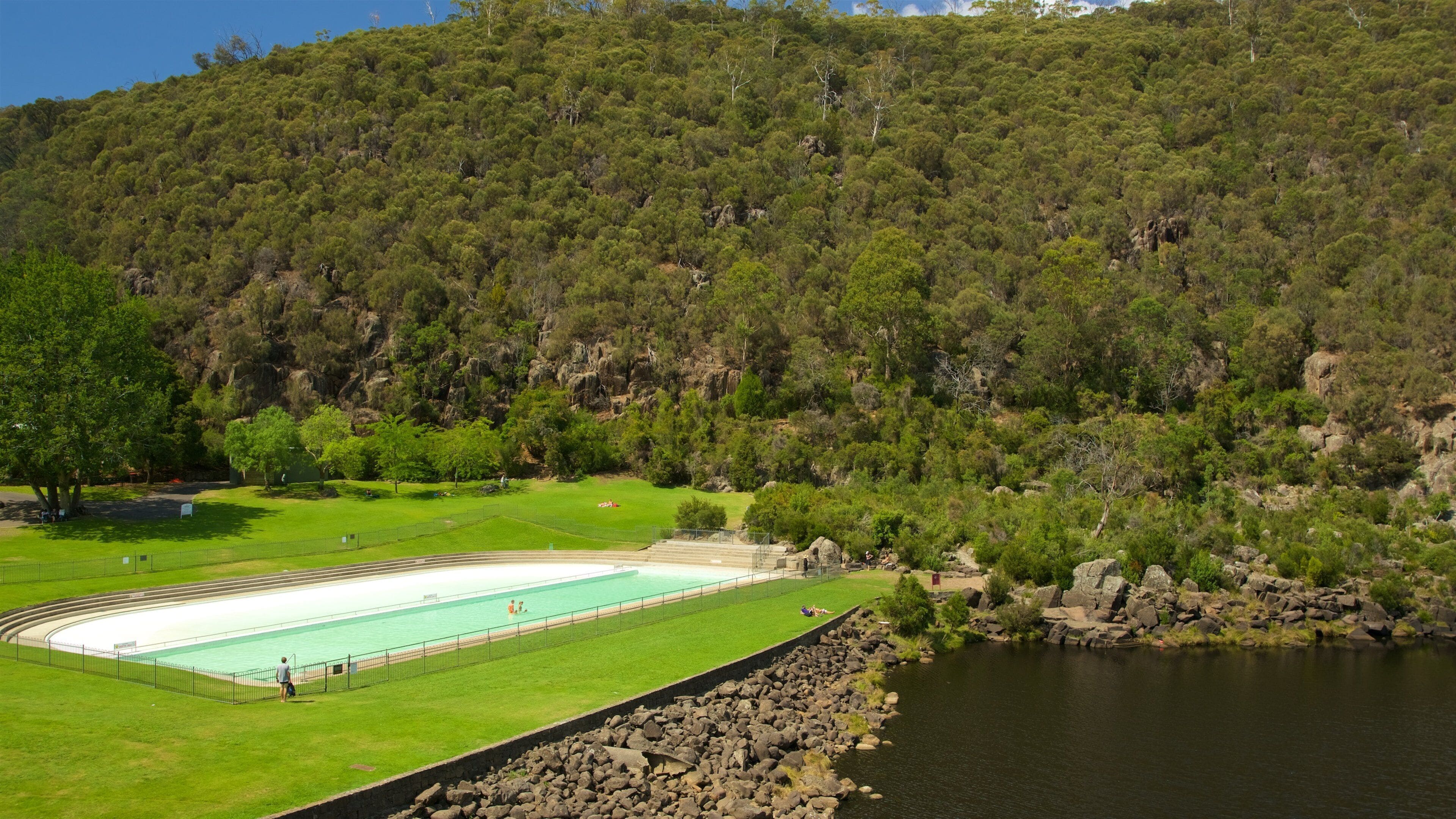 A swimming pool surrounded by a grassy area, a rocky shoreline, and a forested hillside.