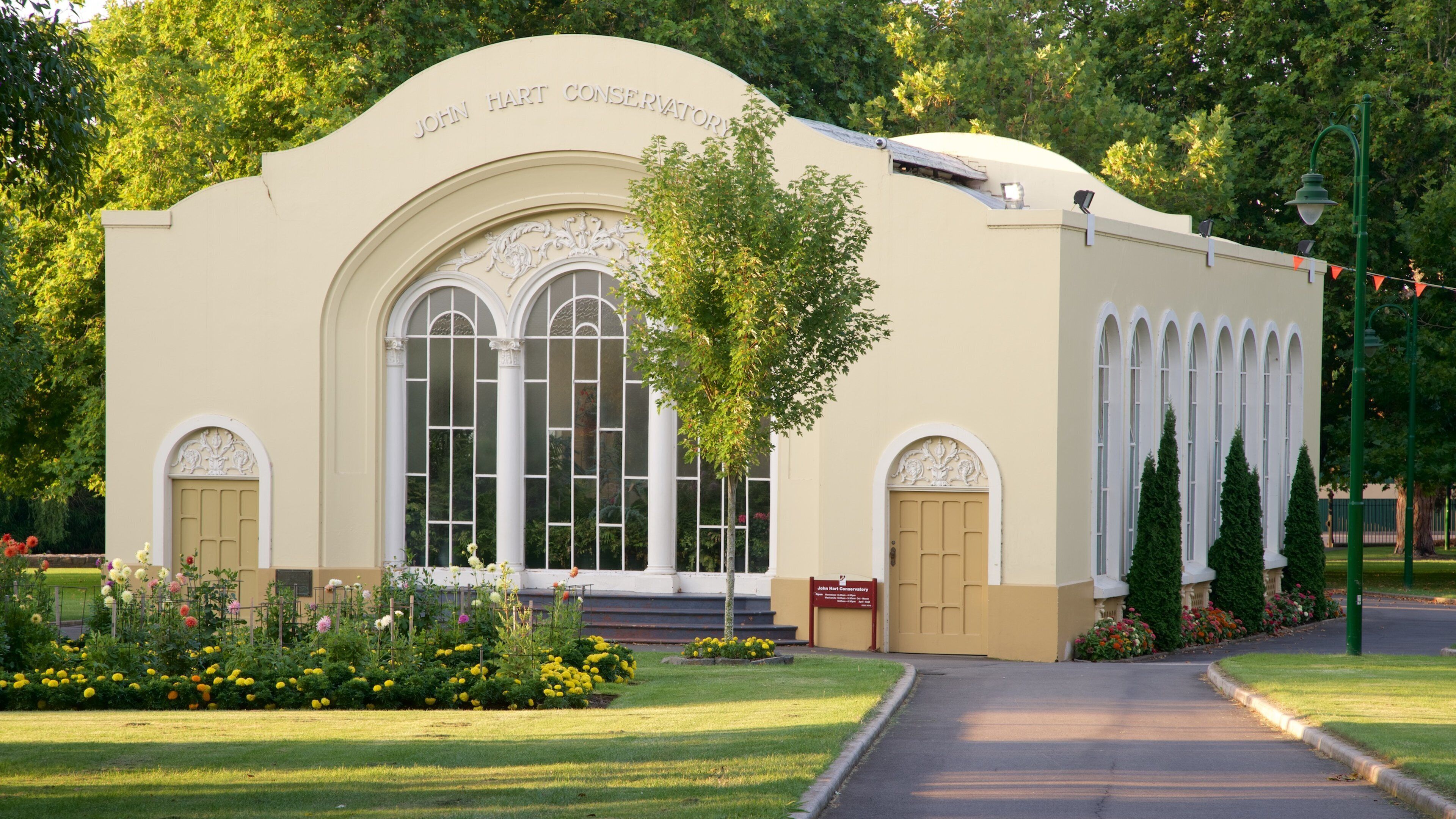 Un invernadero color beige con ventanas arqueadas y un cartel que dice 'John Hart Conservatory'.