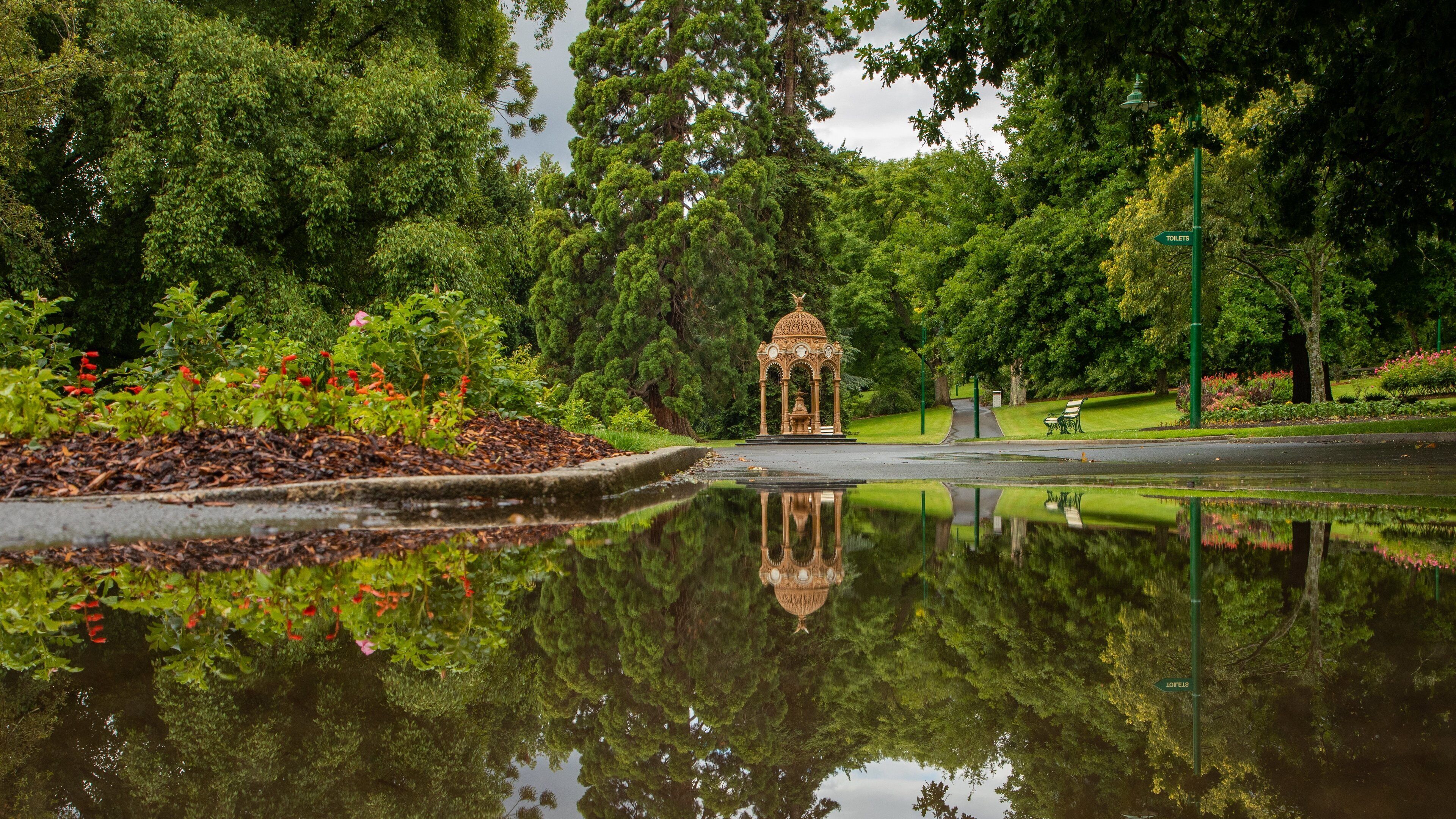 A gazebo in a park with a pond reflecting it, surrounded by lush greenery and a sign indicating toilets.