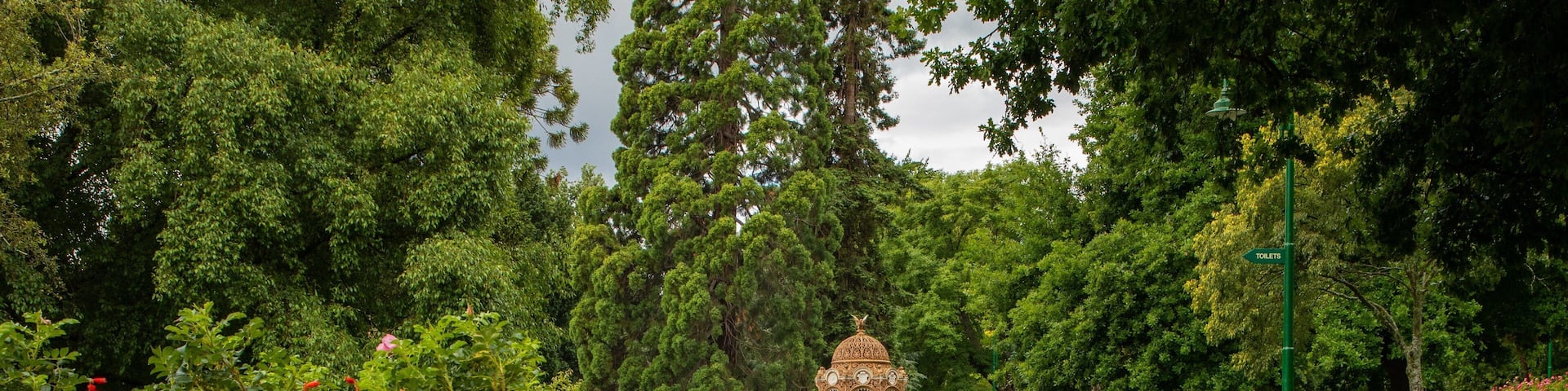 A gazebo in a park with a pond reflecting it, surrounded by lush greenery and a sign indicating toilets.