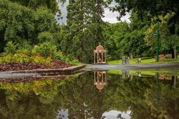 Un kiosque dans un parc, se reflétant dans un étang, entouré d’une végétation luxuriante et agrémenté d’un panneau indiquant l’emplacement des toilettes.
