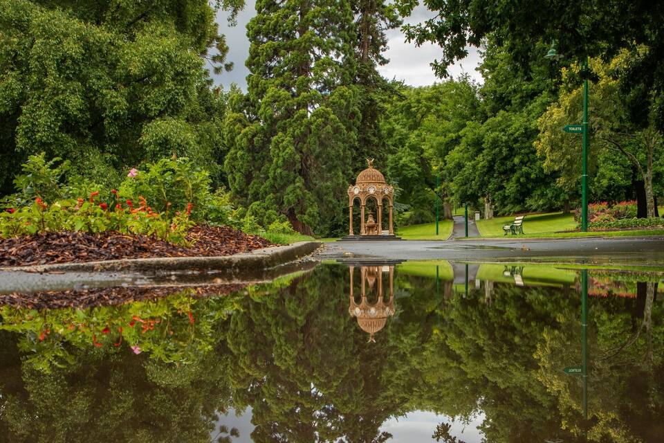 A gazebo in a park with a pond reflecting it, surrounded by lush greenery and a sign indicating toilets.