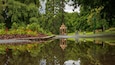 Ein Pavillon in einem Park mit einem Teich, der ihn widerspiegelt, umgeben von üppigem Grün und einem Schild, das Toiletten anzeigt.