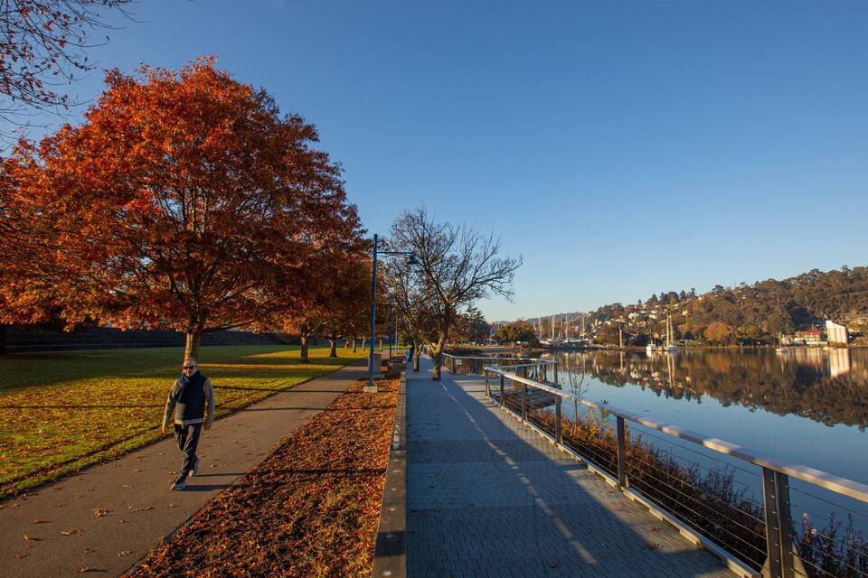 Een voetpad langs de rivier met herfstkleuren, een rustig meer en een stad in de verte.