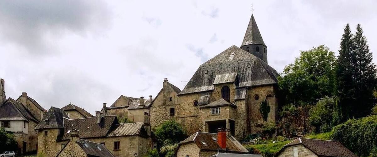 A historic stone village with a prominent church tower, lush greenery, and a flowing river.