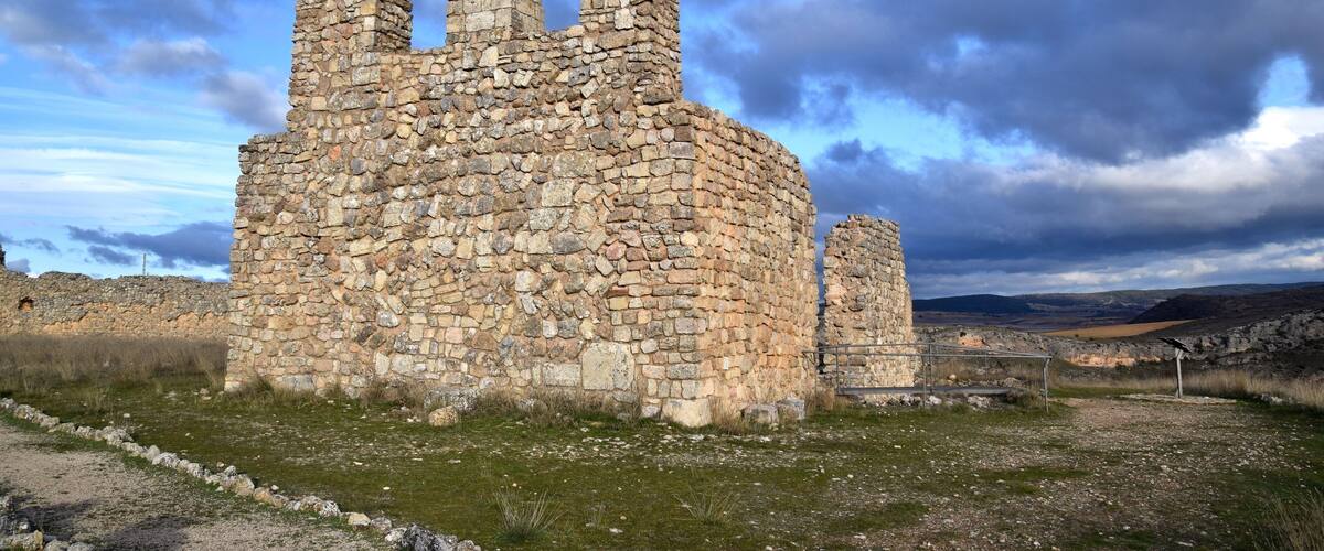 A stone ruin with arched openings and a circular base, surrounded by a grassy field and a cloudy sky.