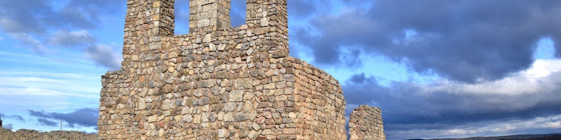 A stone ruin with arched openings and a circular base, surrounded by a grassy field and a cloudy sky.