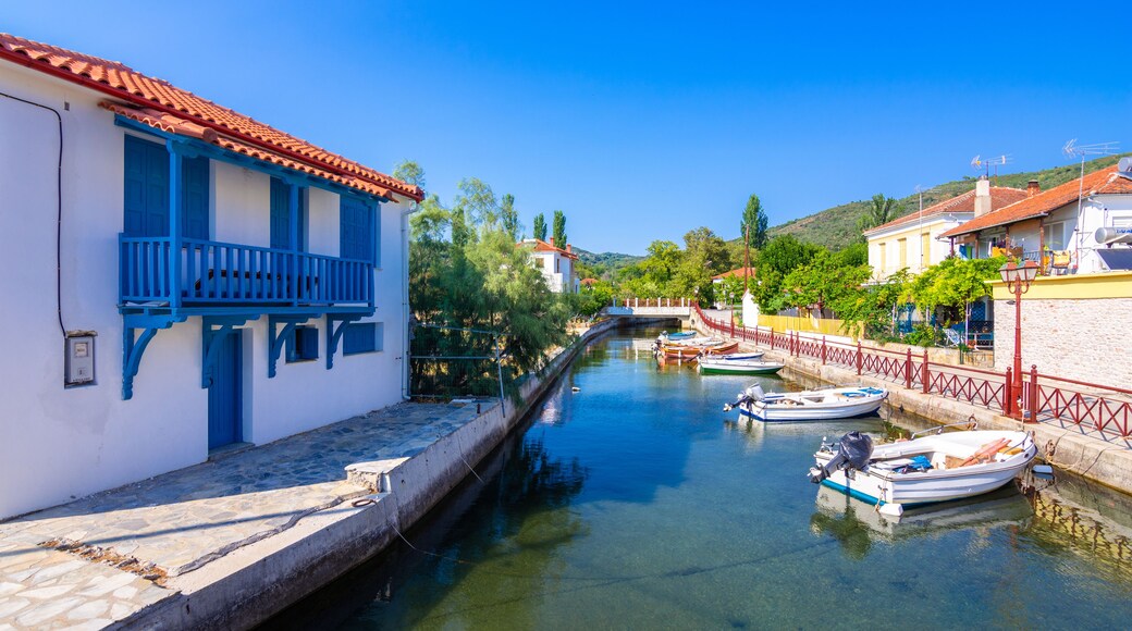 A narrow canal with boats docked on the right, a white building with blue shutters on the left, and a red fence along the canal's edge.