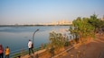 A calm river with a city skyline in the background, a pedestrian walkway with trees, and a person standing by the railing.