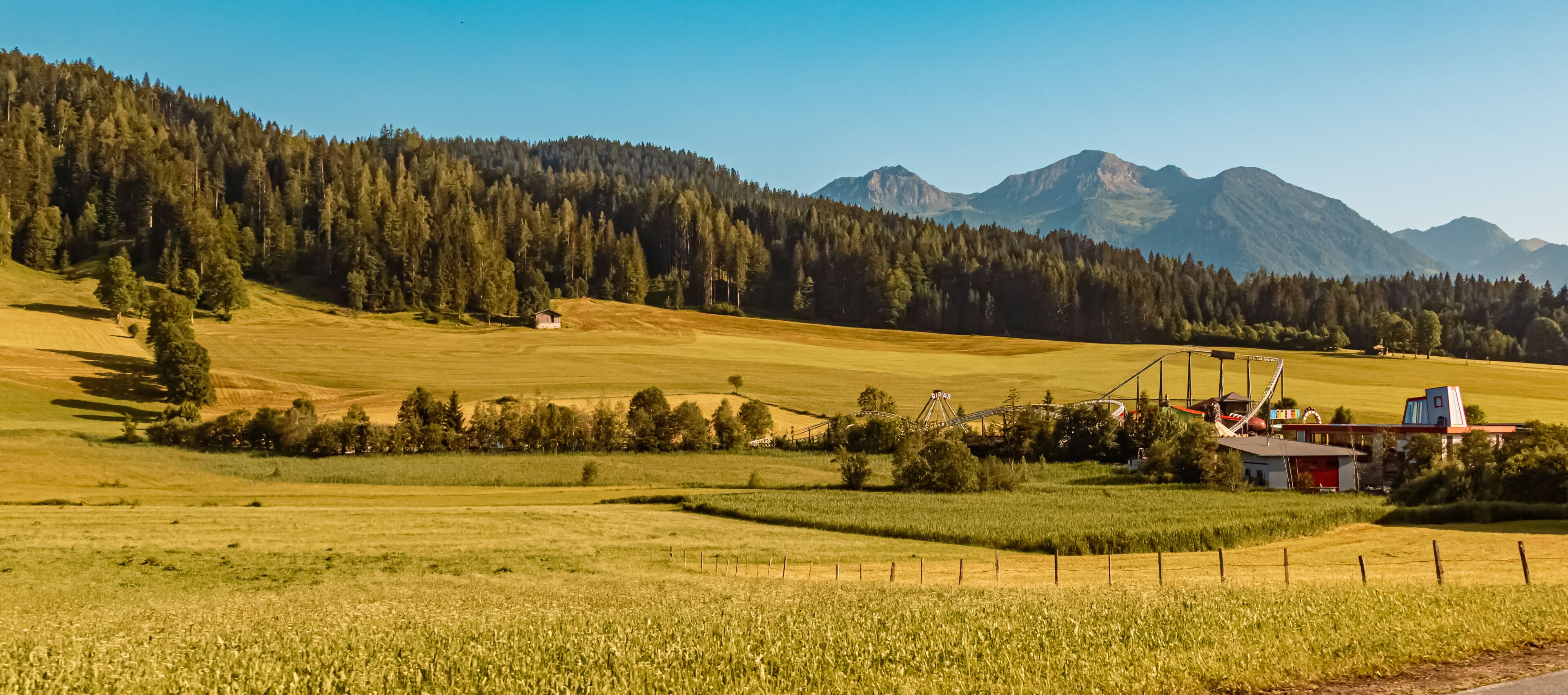 A rural landscape with a field, a small house, and a roller coaster in the background.