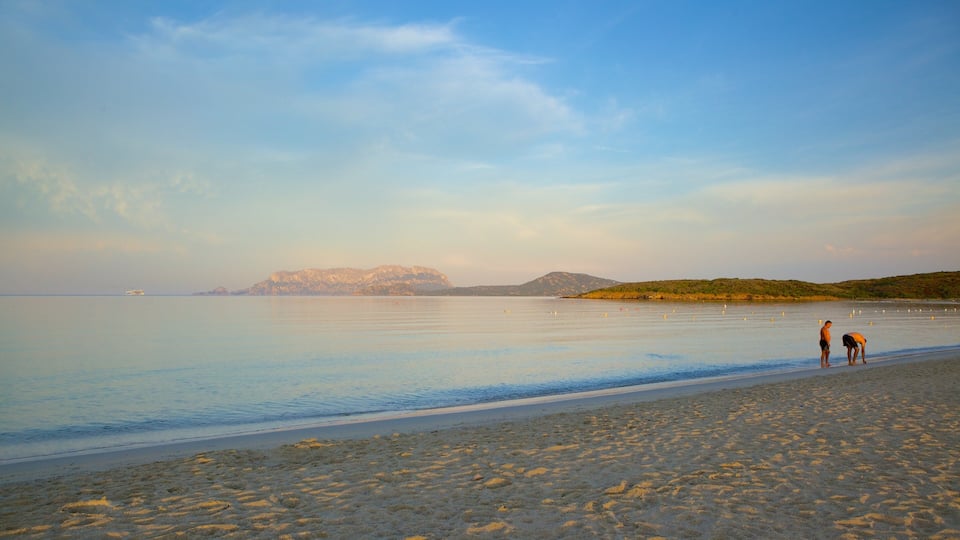 A calm beach with two people standing on the shore, a boat in the distance, and mountains in the background.