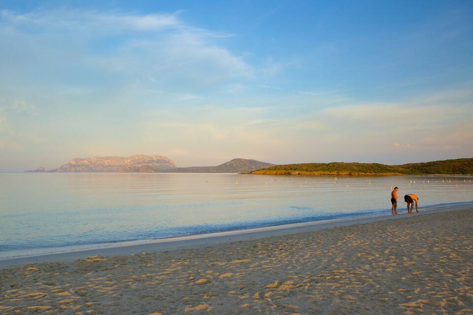 A calm beach with two people standing on the shore, a boat in the distance, and mountains in the background.