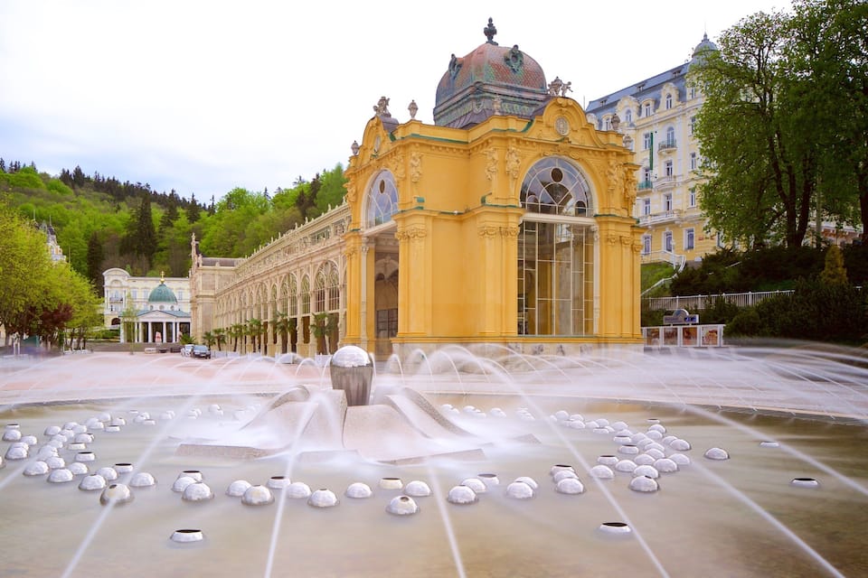 A fountain with water jets in front of a yellow building with ornate architecture.