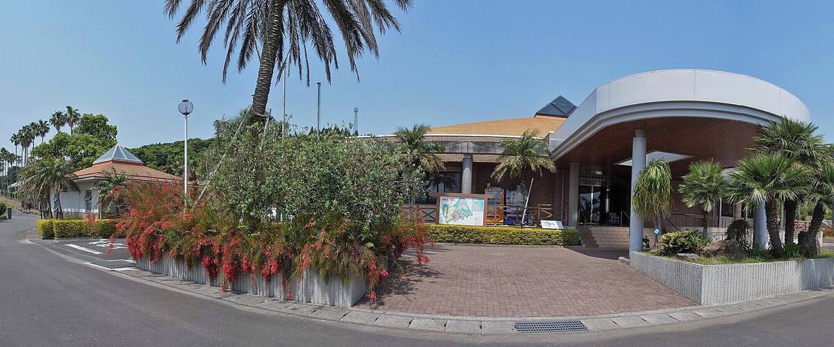 A modern building with a dome and a curved entrance, surrounded by palm trees and a brick pathway.