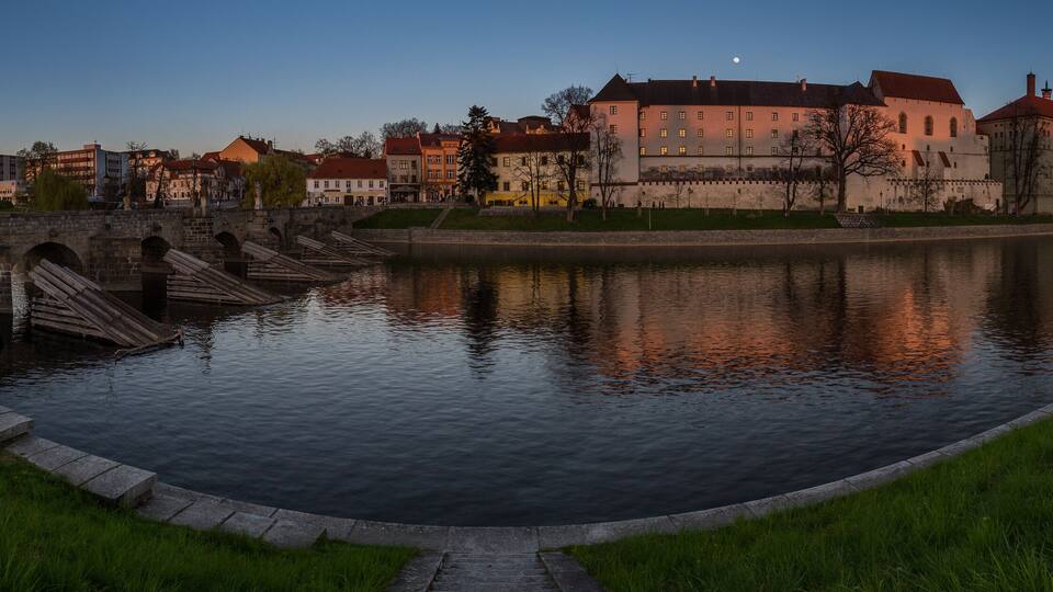 A river with a stone bridge, a castle, and surrounding buildings.