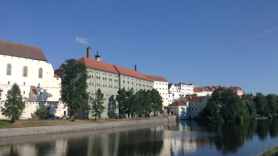 A historic building with a red roof by a calm river, surrounded by trees and other buildings.