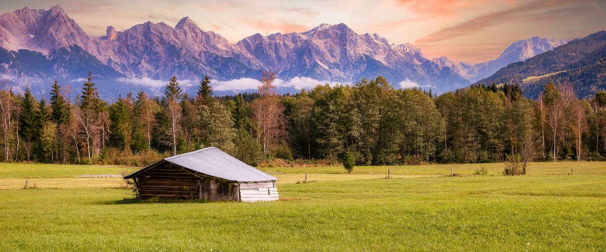 A wooden cabin in a grassy field with a mountain range in the background.