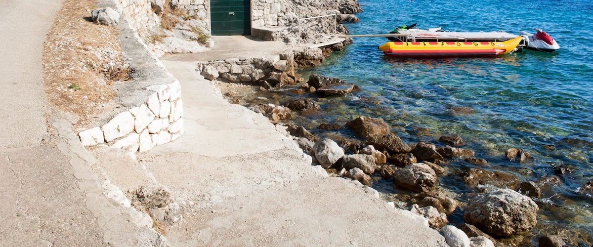 A coastal road with a stone building, clear blue sea, and a yellow boat tied to the shore.