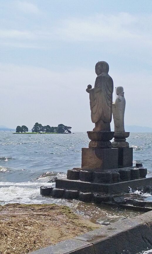 A statue of a figure holding a child, standing on a platform by the sea.