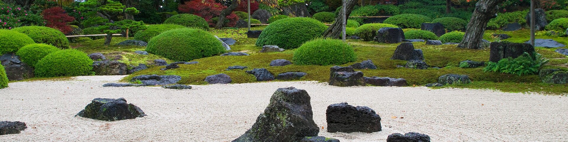 Um jardim japonês sereno com pedras dispostas com cuidado, vegetação exuberante e diversas árvores.
