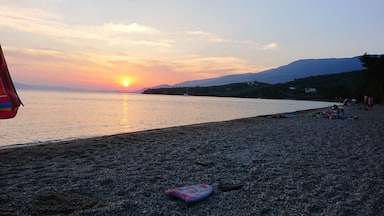 A beach at sunset with the sun setting over the water, a few people sitting on the sand, and mountains in the background.