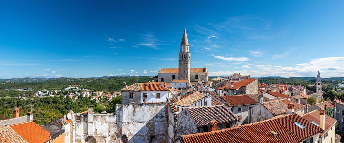 A historic town with a prominent church tower, terracotta-roofed buildings, and a lush green landscape in the background.