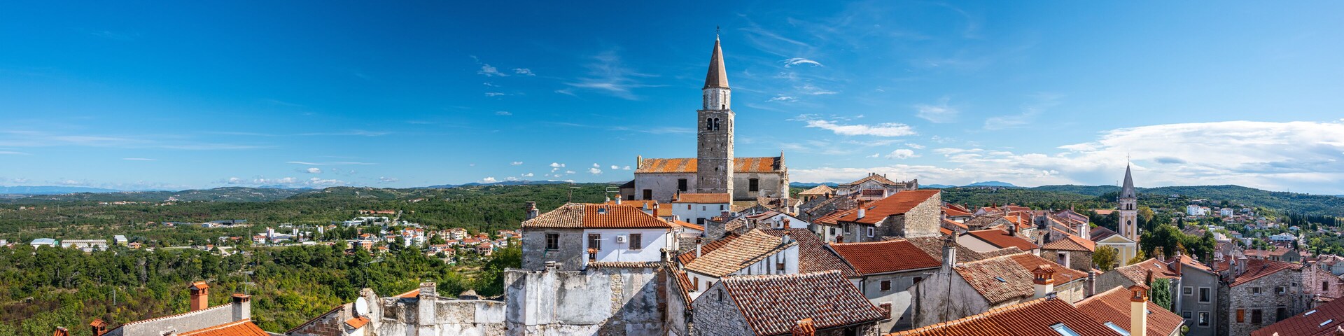 A historic town with a prominent church tower, terracotta-roofed buildings, and a lush green landscape in the background.
